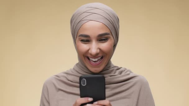 Concepto de ganar en línea. Emocionada joven musulmana leyendo mensaje ganador de correo electrónico en el teléfono celular, fondo beige, cámara lenta — Vídeo de stock
