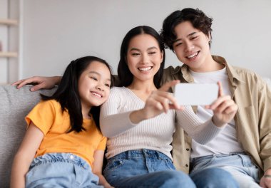 Glad smiling young asian couple and teen daughter making photo on phone on sofa in living room