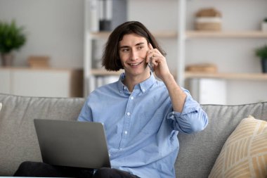 Young guy freelancer having phone conversation while working on laptop from home, looking aside at copy space