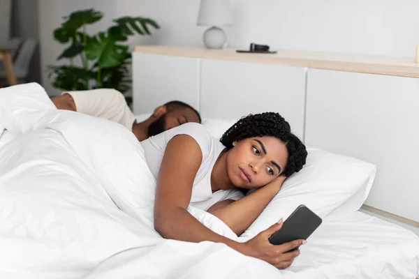 Sad young african american lady looks at smartphone lie on comfortable bed hide cheating — Stock fotografie