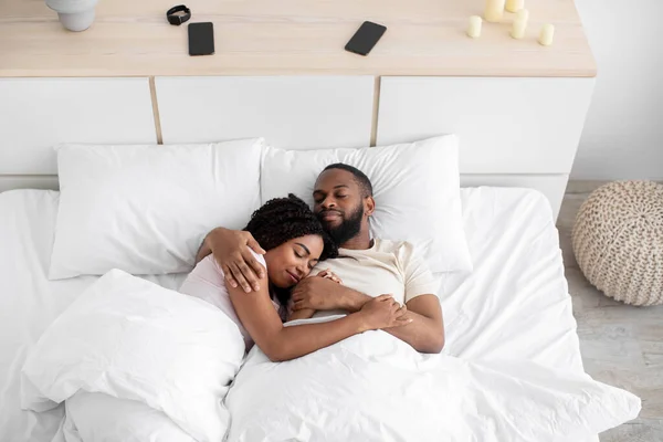 Loving happy peaceful millennial african american man hugs his woman and sleeps on comfortable bed together in bedroom — Stock fotografie