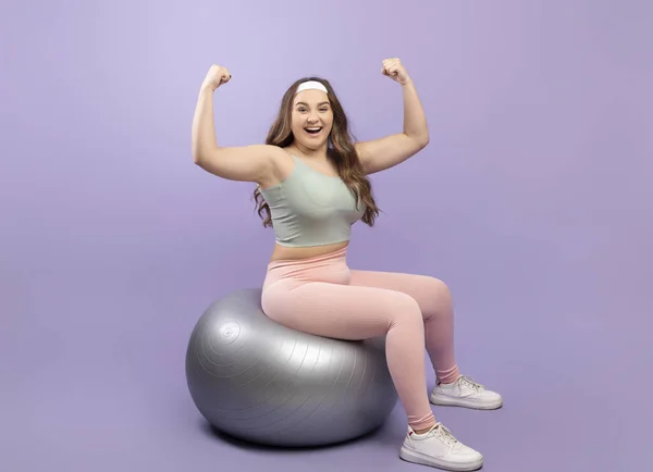 Happy young european plus size woman in sports uniform shows biceps on arms, sits on fitness ball — стокове фото