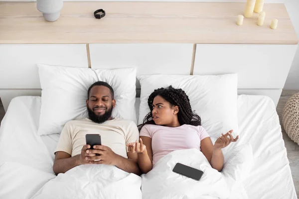 Smiling young african american guy chatting on phone, sad angry offended wife swears on comfortable bed — 스톡 사진
