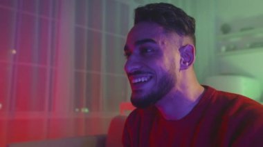 Young emotional middle eastern guy watching exciting tv program or playing video games, sitting at home in neon lights
