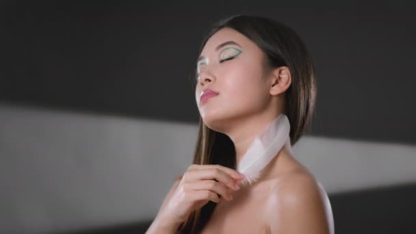 Vücut bakımı prosedürleri. Sanatı olan genç, bakımlı Asyalı kadın pürüzsüz tenini beyaz tüyle okşuyor. — Stok video
