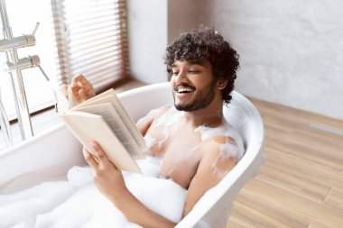 Spa prosedürü, kişisel bakım ve banyoda dinlenme. Köpüklü mutlu Hintli genç adam, sabahları kitap okuyor.