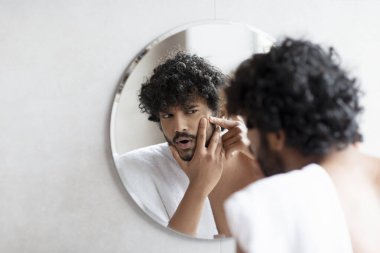 Problem skin. Concerned indian man popping pimple on cheek while standing near mirror in bathroom