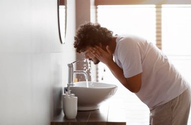 Young indian man washing face with clear water from tap bending over sink, doing skincare facial routine in bathroom