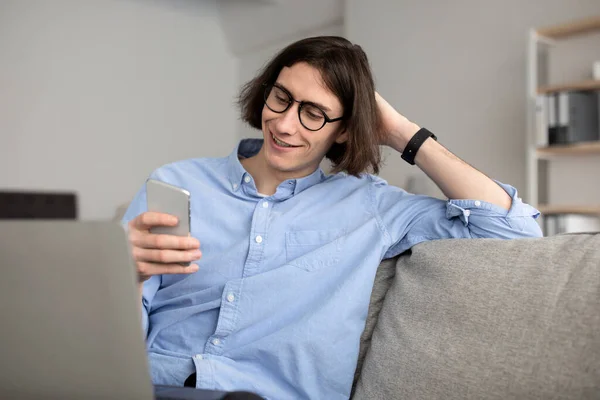 Happy young guy enjoying video chat with lover, looking at smartphone screen, resting while working on laptop — Photo