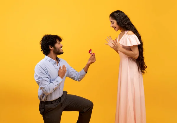 Loving indian man standing on one knee and offering engagement ring to his beloved woman on yellow background — Zdjęcie stockowe