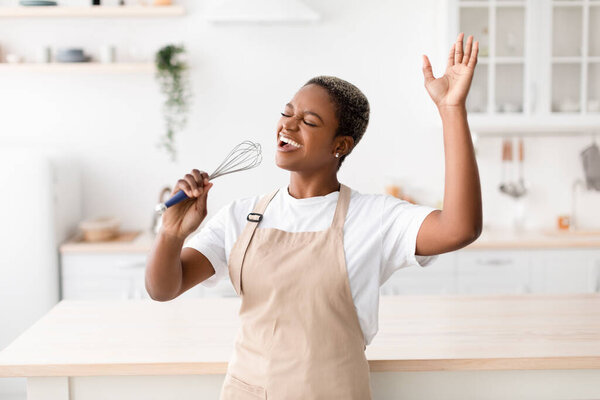 Happy inspired young african american woman in apron prepares food, sings at imaginary microphone