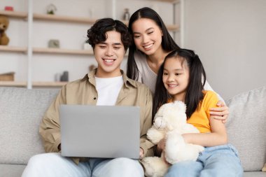 Smiling cheerful chinese young couple and girl look at laptop for ad and offer, sitting on sofa in living room