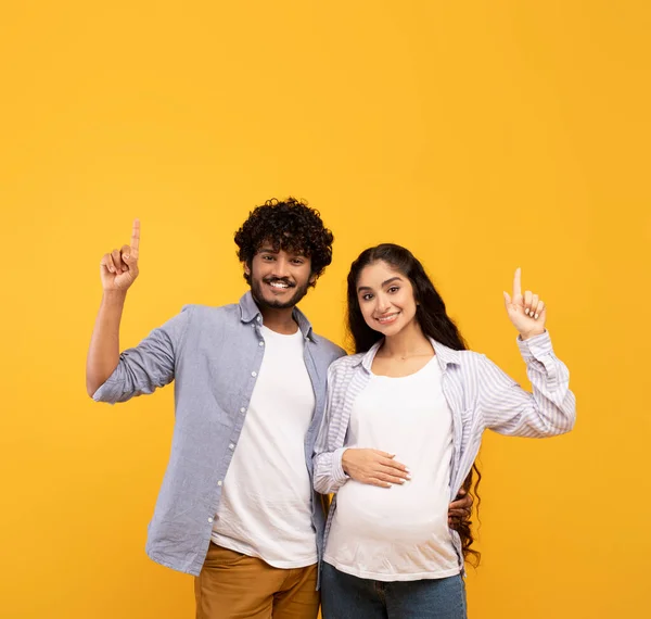 Excited indian pregnant lady and her husband pointing up at free space, showing amazing sale or offer, yellow background — ストック写真