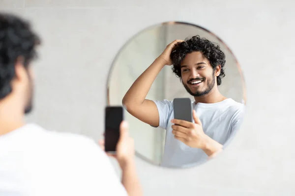 Cheerful indian man standing next to mirror in modern bathroom, holding smartphone while doing morning routine — Stockfoto