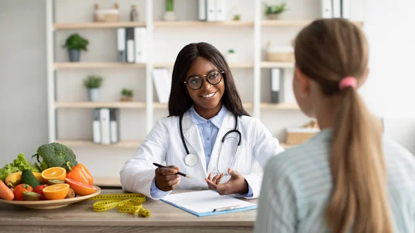 Amigável afro-americano nutricionista feminino dando consulta ao paciente em sua clínica, discutindo diário de dieta — Fotografia de Stock
