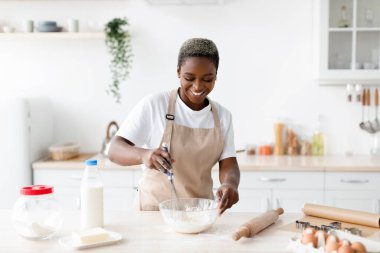 Glad happy young african american female in apron make dough for baking, prepares eat in scandinavian kitchen