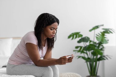Despaired sad millennial african american lady sit on bed and holding pregnancy test in bedroom interior, profile