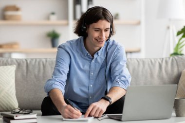 Çevrimiçi eğitim kavramı. Kulaklıklı genç adam evde dizüstü bilgisayarla çalışıyor, Webinar izlerken notlar alıyor.