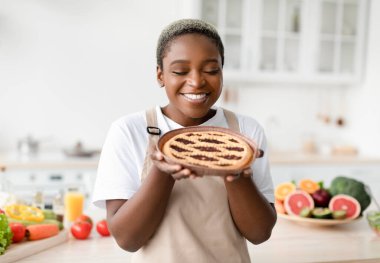 Smiling pretty young african american woman in apron sniffs smell of fresh pie in scandinavian kitchen interior