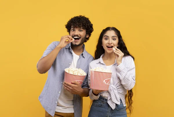 Excited indian man and woman eating popcorn at cinema, standing over yellow studio background — Stockfoto