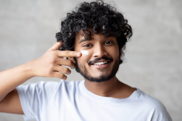 Handsome indian man applying moisturising cream on face while standing in bathroom, doing morning routine — Φωτογραφία Αρχείου