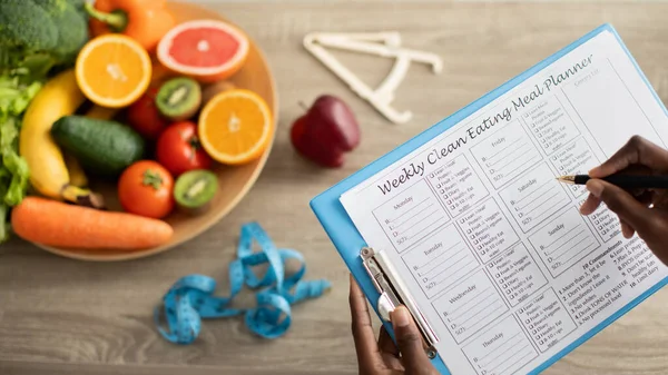 African american nutritionist doctor writing weekly diet plan, sitting at desk with plate of fruits and vegetables — Foto Stock