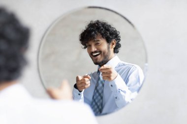 Youre handsome. Confident indian businessman pointing at mirror in bathroom with two fingers, wearing shirt and smiling