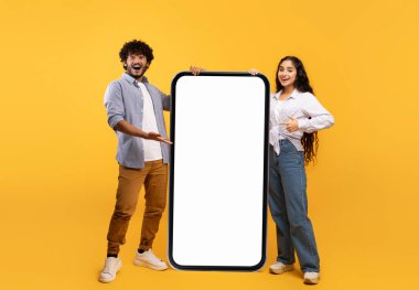 Güzel cep telefonu teklifi. Mutlu Hintli çift, beyaz ekranlı, sarı arka planlı, büyük cep telefonuna bakıyor.
