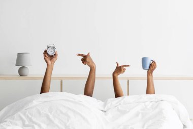 Hands of young black guy and woman stick out from under blankets, holds cups and point finger at alarm clock