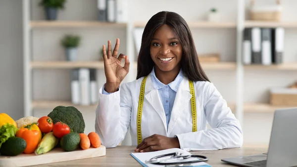African american female nutritionist showing okay gesture, sitting at table with laptop and fresh products in clinic —  Fotos de Stock
