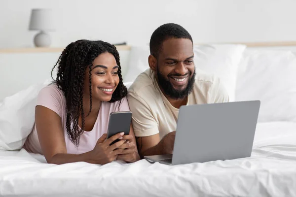 Χαρούμενος νεαρός αφροαμερικάνος σύζυγος και σύζυγος με smartphone, ξαπλώνουν σε άνετο κρεβάτι και κοιτάζουν το laptop — Φωτογραφία Αρχείου