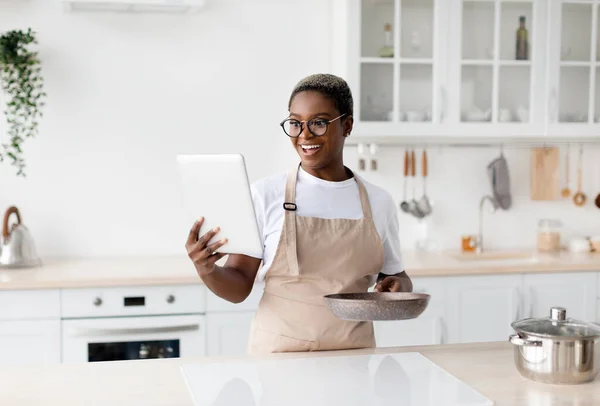 Surprised happy young african american woman in apron and glasses looks at tablet and prepares dinner — 스톡 사진