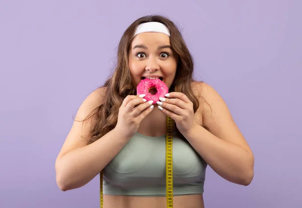 Portrait of smiling hungry funny millennial european woman plus size in sports uniform with measuring tape eating donut — Stock fotografie