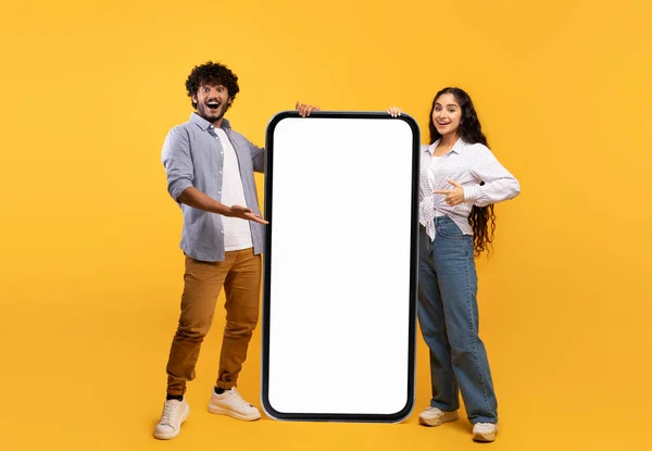 Cool mobile offer. Happy indian couple pointing at big cellphone with white screen for mockup, yellow background — Stockfoto