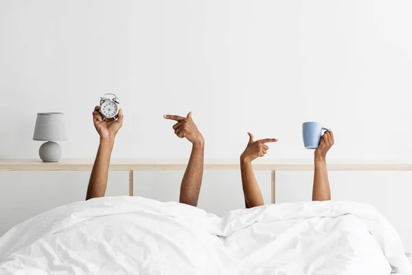 Hands of young black guy and woman stick out from under blankets, holds cups and point finger at alarm clock — ストック写真