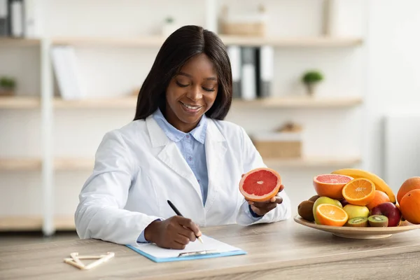 Consejera de nutrición femenina negra feliz sosteniendo pomelo, haciendo plan de comidas para el cliente en la clínica de pérdida de peso —  Fotos de Stock