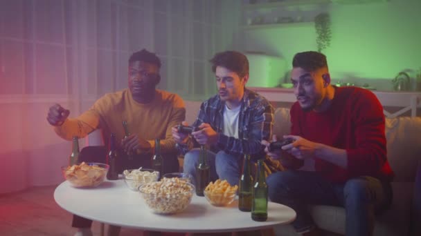 Joven alegre diversos amigos masculinos jugando videojuegos con joysticks, descansando en casa por la noche con cerveza y aperitivos — Vídeo de stock