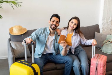 New trip, finally. Excited young tourists couple holding tickets and passports, sitting with packed suitcases on sofa