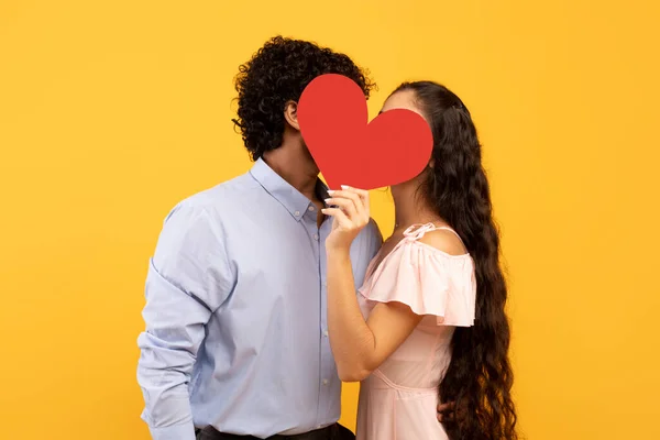 Young indian sweethearts hiding behind red paper heart and kissing, covering their faces, yellow background, side view — Stock Fotó