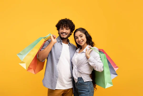 Big sale and shopping with pleasure. Happy young indian couple holding many colored packages in hands, yellow background — Stok fotoğraf