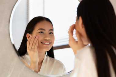 Mirror reflection of happy asian woman applying eye care product