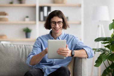 Excited young man chilling at home with digital tablet, sitting on sofa and surfing internet, copy space