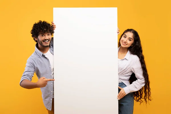 Happy indian couple in love standing by white empty board for advertisement or text over yellow studio background — Stok fotoğraf