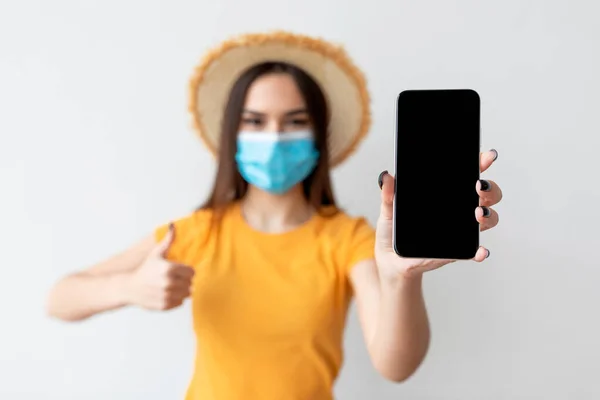 Young woman wearing protective face mask and hat, showing smartphone with empty blank screen on light wall — Stock Photo, Image