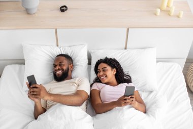 Happy laughing young african american wife and husband with smartphones lies on comfortable bed in bedroom at free time