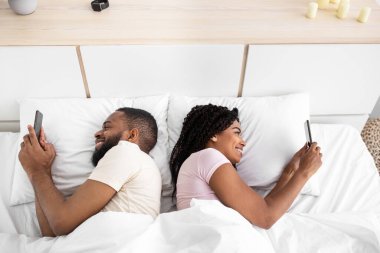 Glad young african american guy and lady chatting on smartphones lie on comfortable bed in bedroom