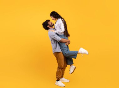 Loving indian guy lifting his girlfriend and holding her on yellow studio background, profile side view, free space banner