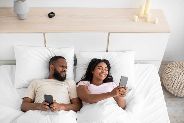 Feliz jovem Africano americano esposa conversando no smartphone, triste marido ofendido olha para gadget — Fotografia de Stock