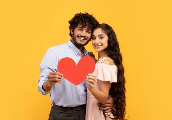 Concept de Saint-Valentin. Portrait de couple indien amoureux tenant coeur de papier rouge et embrassant, fond jaune — Photo