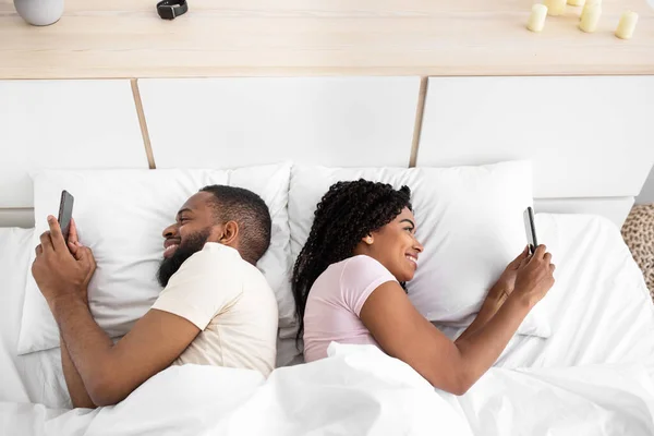 Glad young african american guy and lady chatting on smartphones lie on comfortable bed in bedroom — Fotografia de Stock